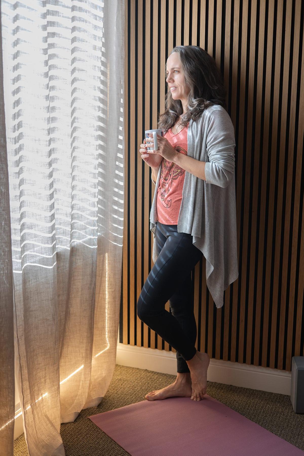 Branding photo of a yoga teacher holding a cup of tea by a sunlit window in her studio