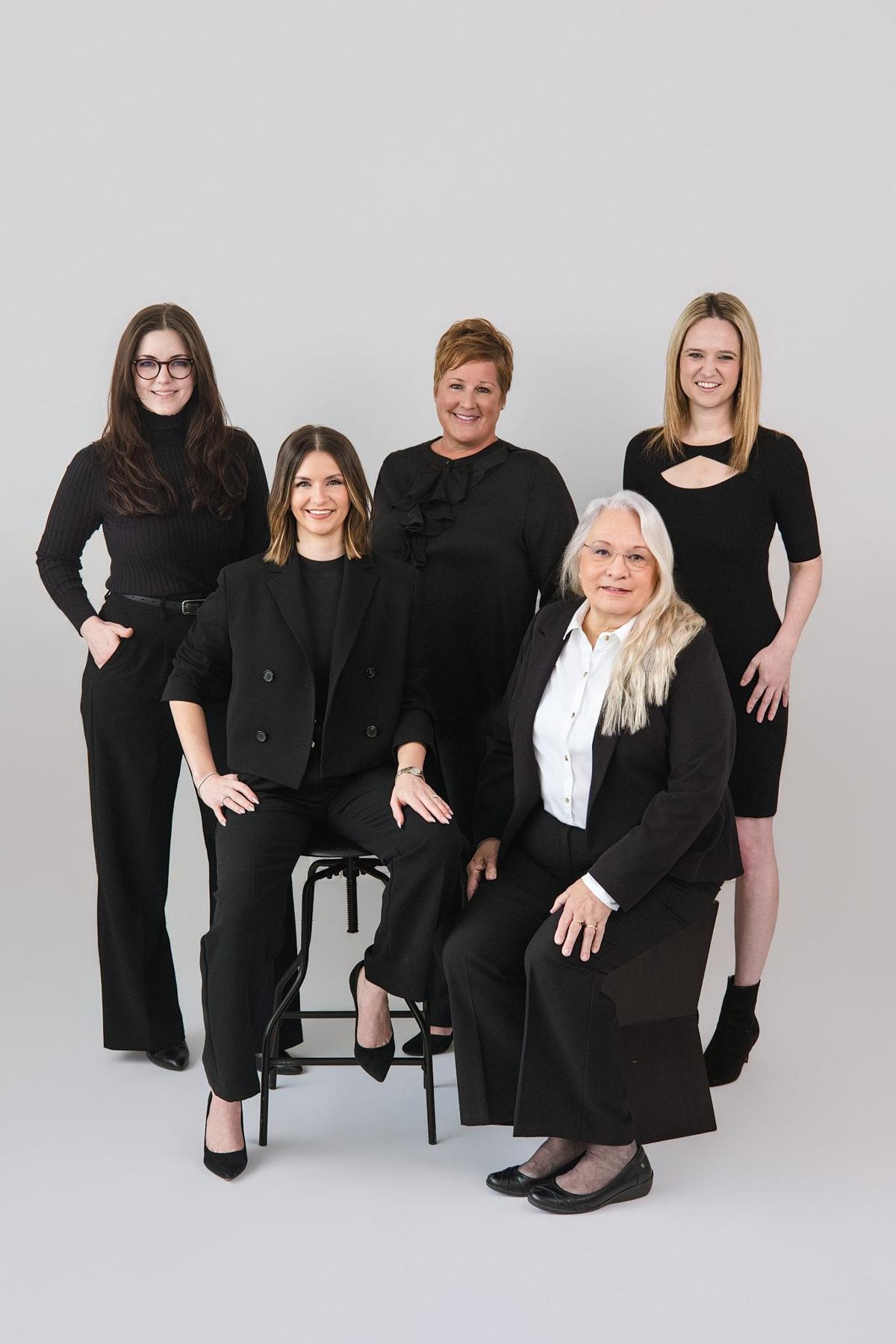 Team photo of five women dressed in black posed together in a white studio