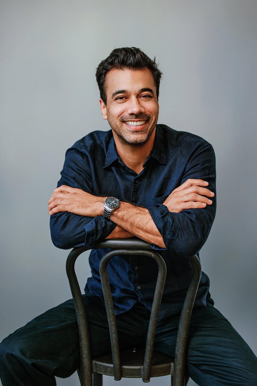 Studio headshot of a smiling man in a navy shirt, arms crossed over the back of a chair